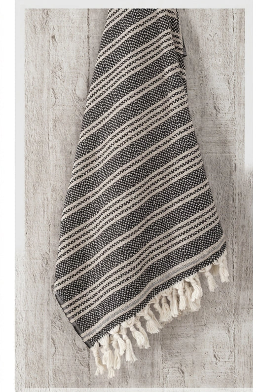 Black and ivory Mevsim Turkish peshtemal towel hanging against a textured wall, showing its woven stripe design and cotton tassels.