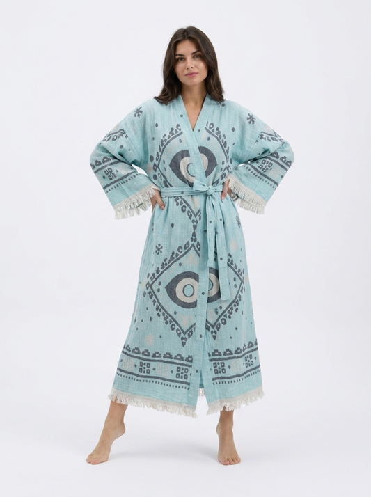Front view of a model wearing a long aqua blue cotton kimono robe with oversized charcoal and cream evil eye motifs, geometric border details, matching waist belt, wide sleeves, and cream fringe trim.