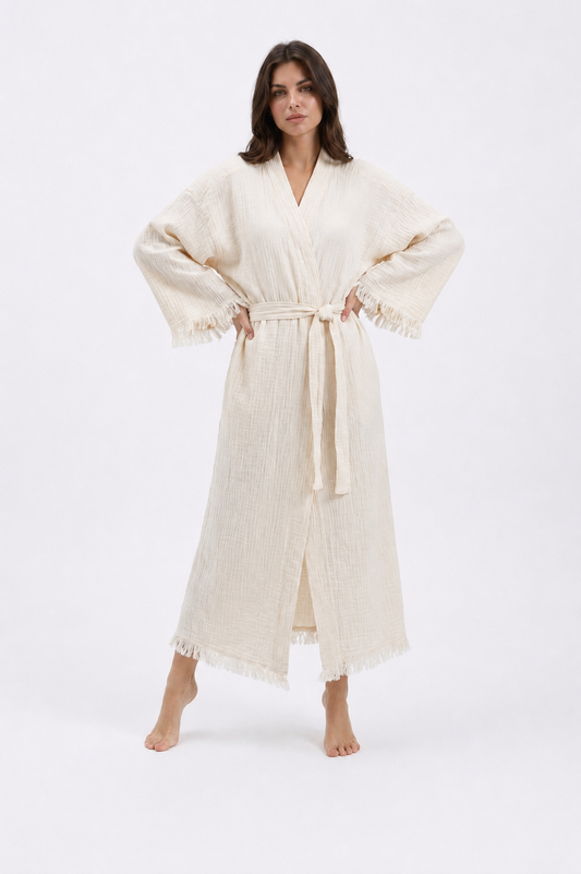 Front view of a model wearing a cream natural cotton kimono robe with a wrap neckline, matching tie belt, wide sleeves, soft fringe trim, and a relaxed ankle-length fit.