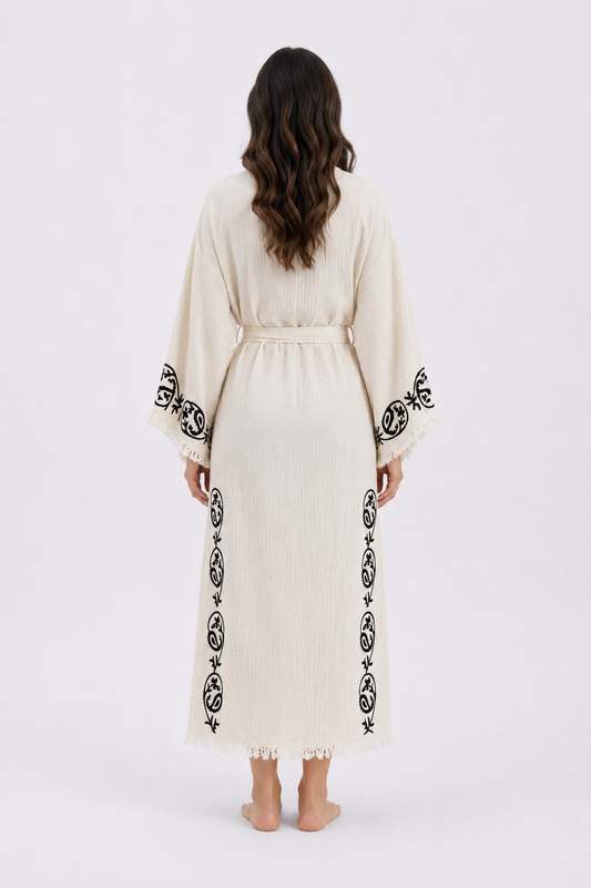 Back view of a long cream cotton kimono robe with handmade black wood block printed botanical motifs along the wide sleeves and lower sides, finished with a matching waist belt, side slits, textured fabric, and delicate fringe trim.