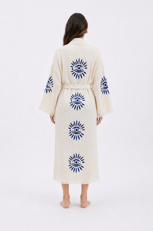 Back view of a cream cotton kimono robe featuring cobalt blue evil eye motifs arranged vertically across the back and sleeves, with a tied waist, relaxed fit, wide sleeves, and soft fringed edges.