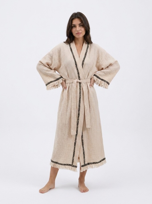Front view of a model wearing a cream textured cotton kimono robe with black patterned trim, a matching waist belt, wide sleeves, fringe cuffs, and a relaxed ankle-length fit.