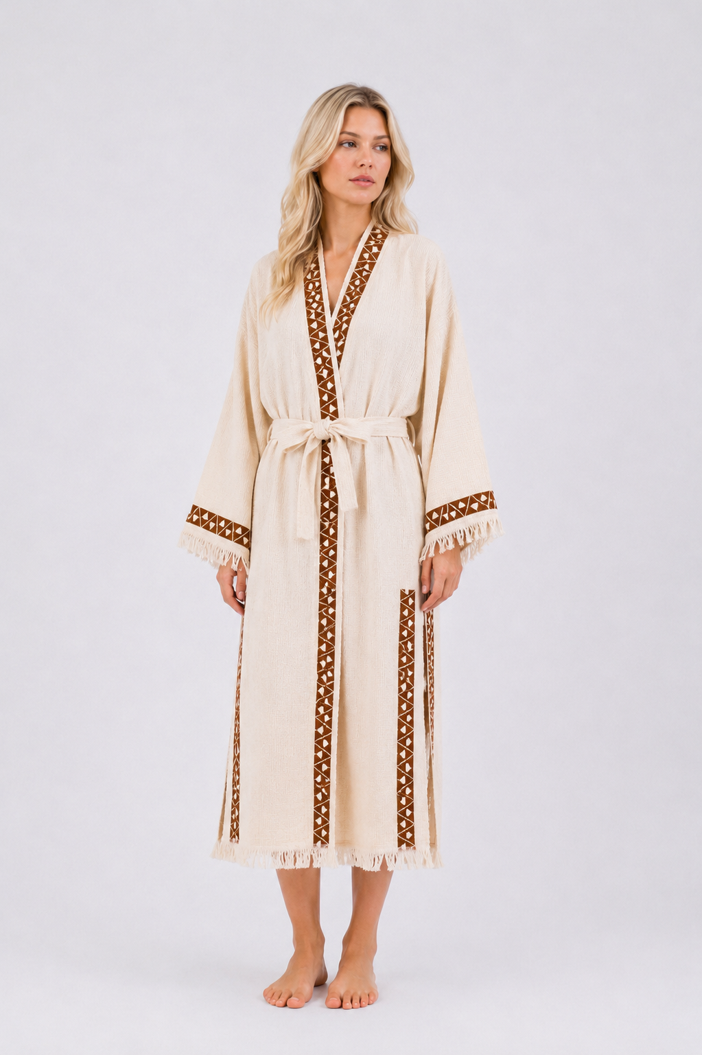 Front view of a model wearing a long cream cotton kimono robe with brown geometric neckline and cuff trim, matching waist belt, wide fringed sleeves, side slits, and an ankle-length hem.
