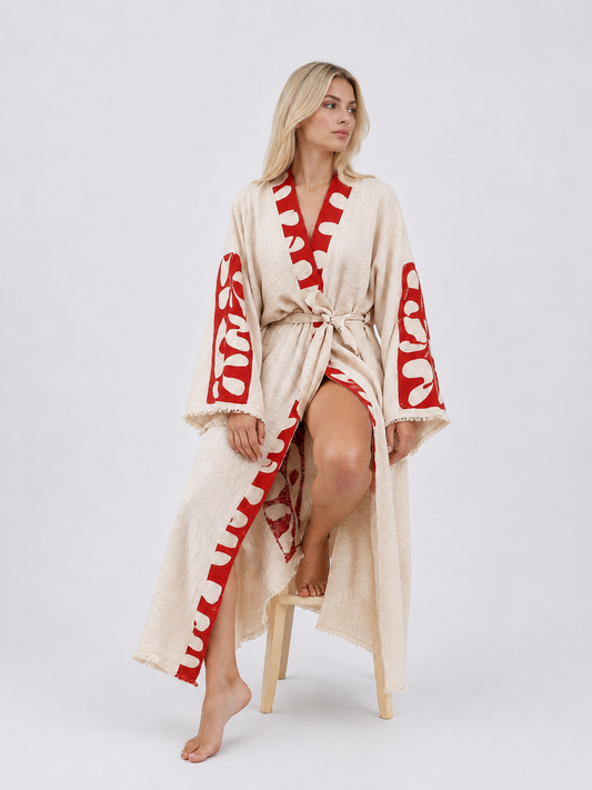 Seated model wearing a long cream textured cotton kimono robe with bold red abstract panels, wide fringed sleeves, a matching waist belt, and a relaxed flowing silhouette.