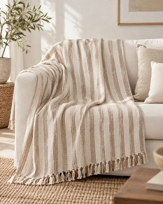 Gelin large cotton throw blanket draped over a cream sofa, showing its natural ivory and taupe striped weave and tassels.