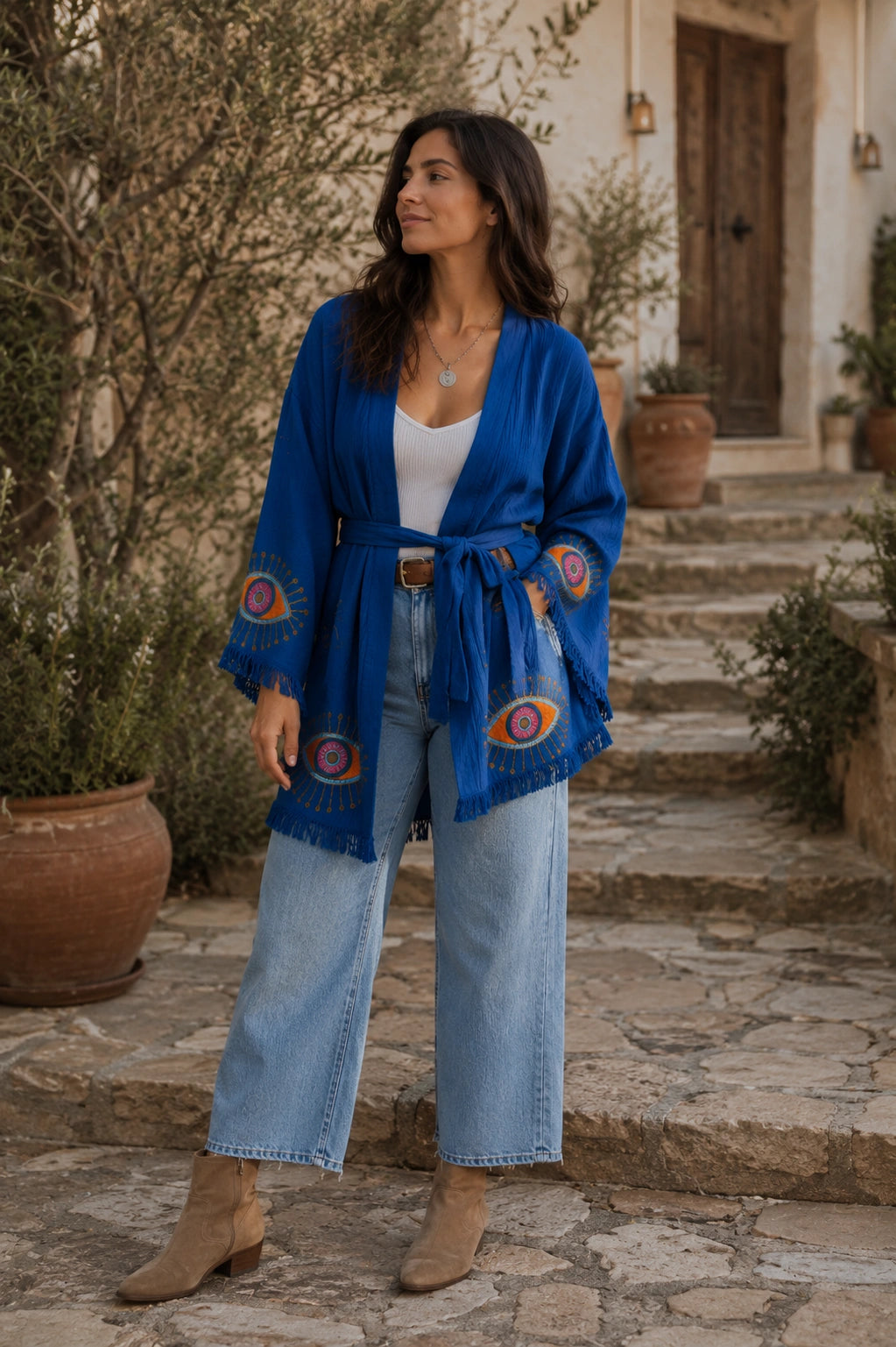 Woman styling the Zaria Blue Protection Kimono top with jeans and boots, showing the versatile cobalt blue wrap jacket with fringe trim and colorful protective eye motifs.