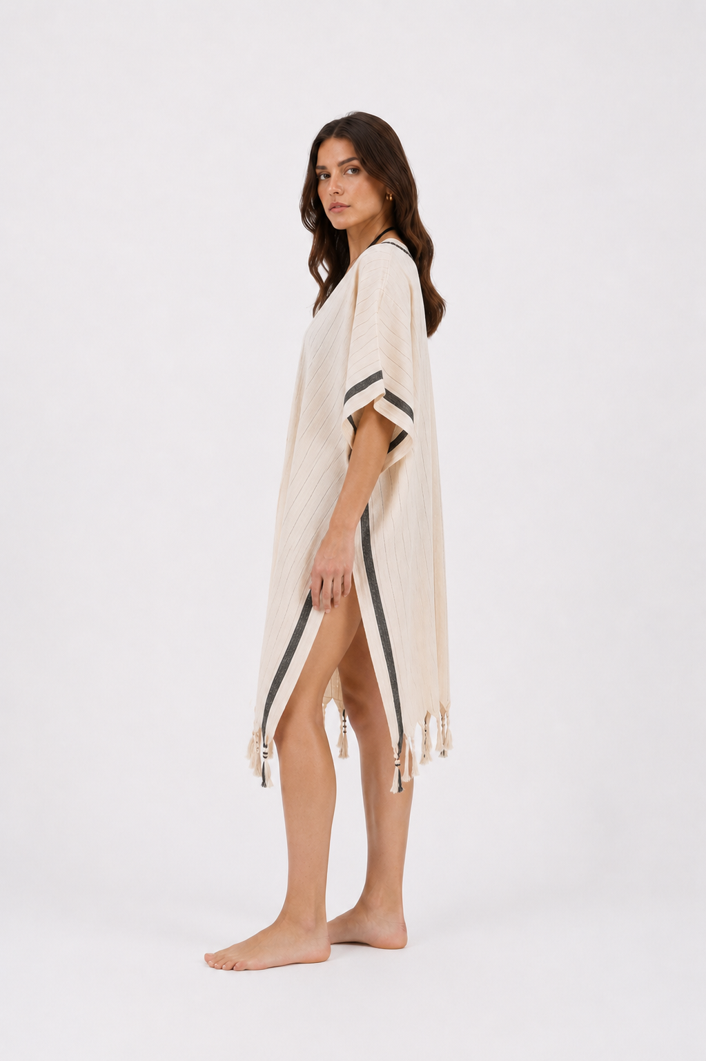 A woman modeling a cream-colored cotton cover-up with black stripe details and tassels, standing against a plain white studio background.