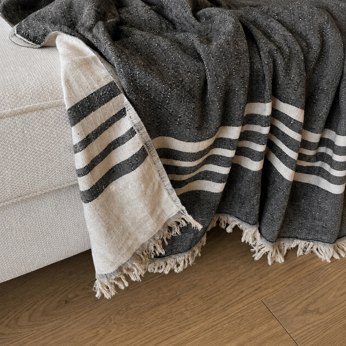 Ela black and natural linen cotton throw blanket draped over a cream sofa showing woven stripes, natural reverse and fringed edges.