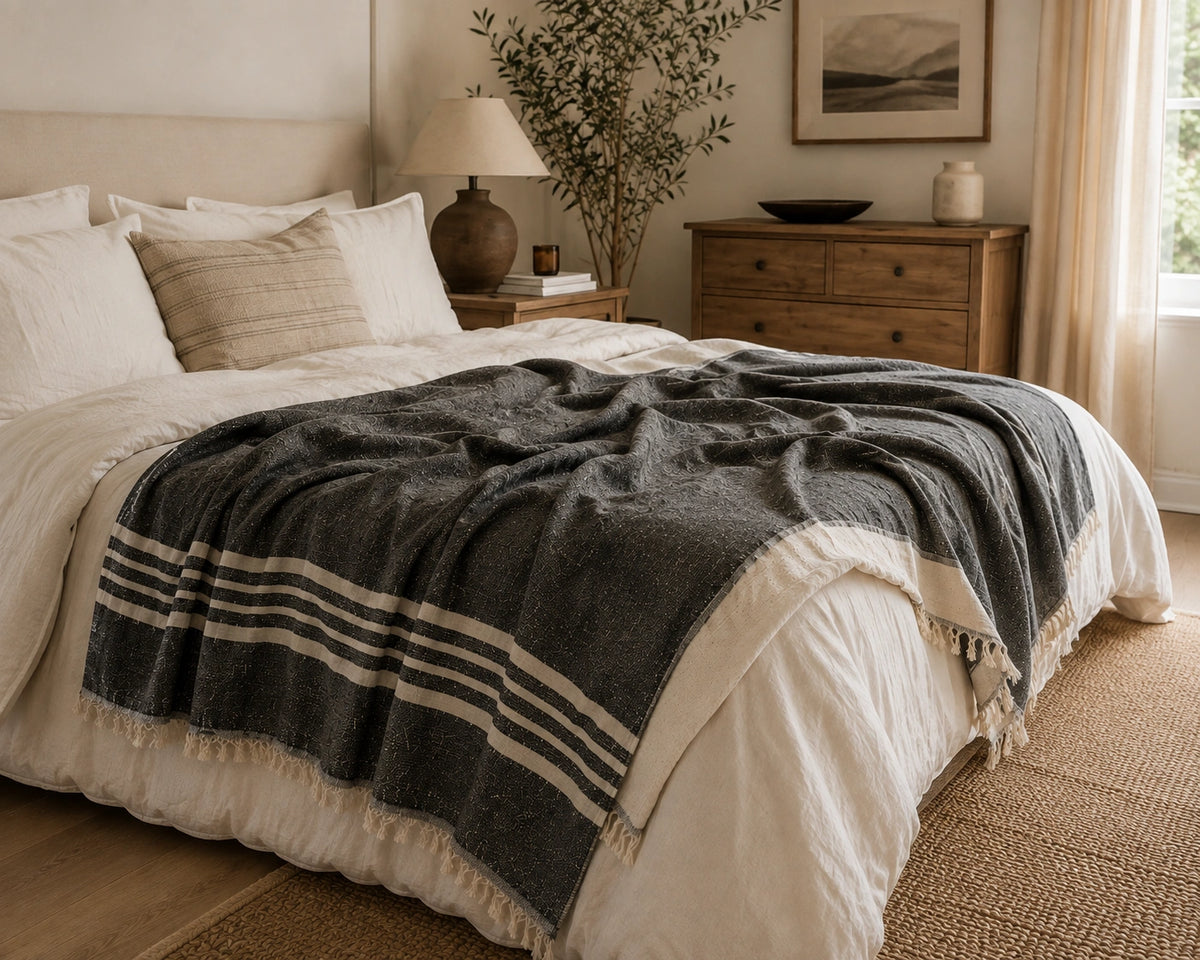 Ela black and natural striped linen cotton throw blanket layered across a neutral bed with fringed edges.