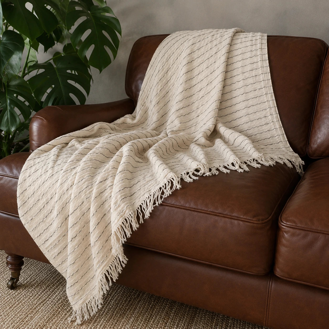 Heidi Turkish cotton throw blanket draped over a brown leather sofa, showing its natural striped weave and fringed edges.