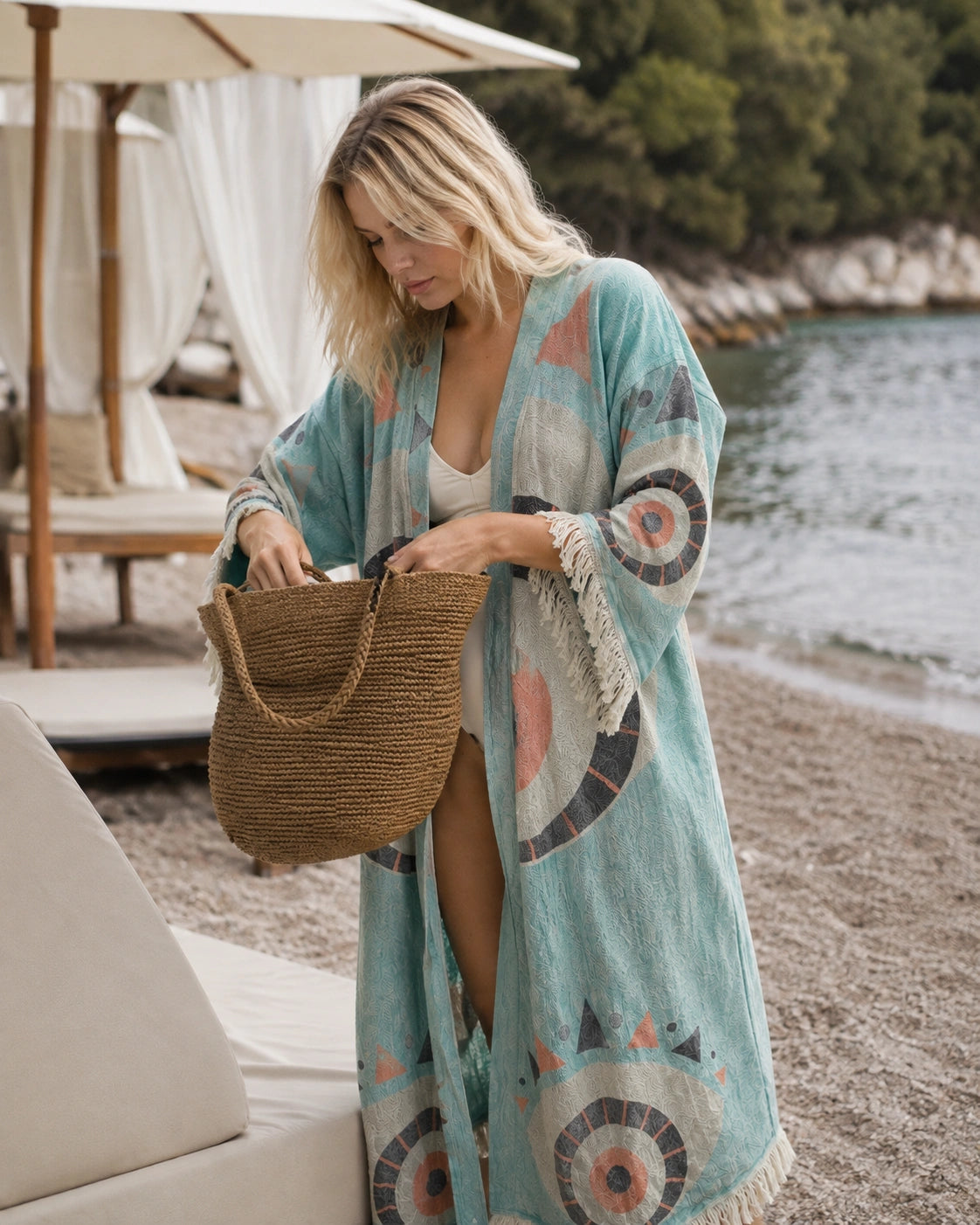 A woman on a beach wearing Liam kimono as a  cover-up with circular patterns and holding a woven straw tote bag.