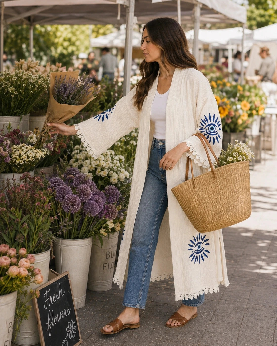 A woman stands at an outdoor flower market wearing a long cream open-front Nazar kimono with blue sunburst embroidery, blue jeans, and brown sandals.