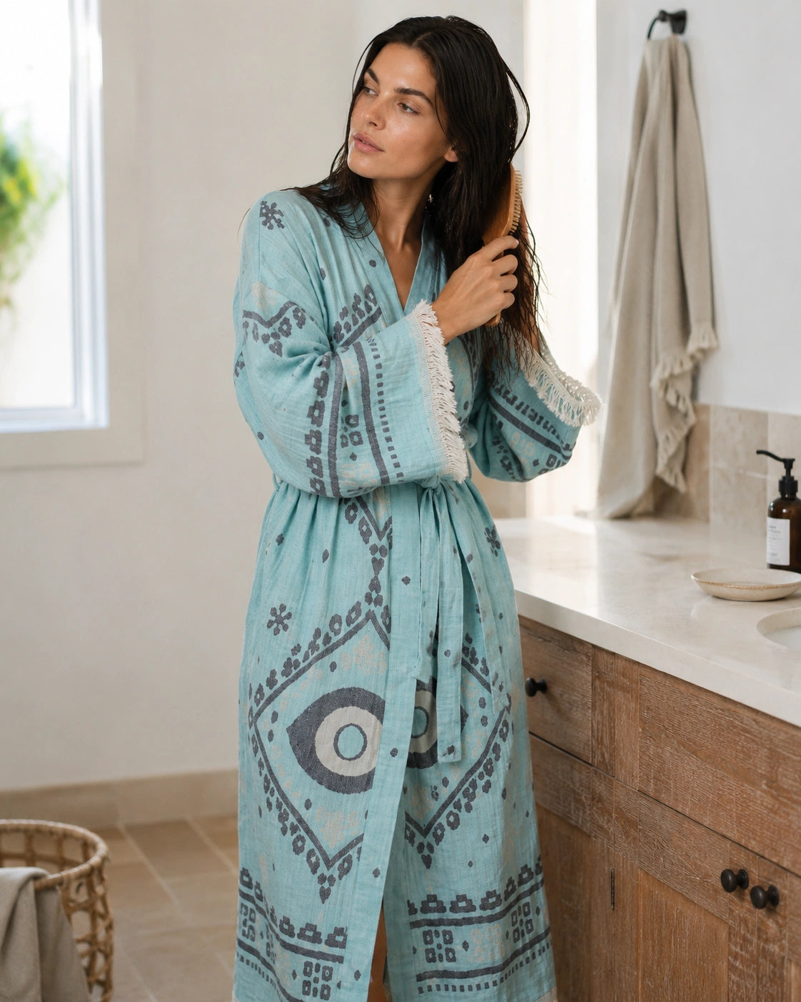 Woman wearing the blue Ocean cotton kimono as an after-shower bathrobe, brushing wet hair in a bright natural bathroom.