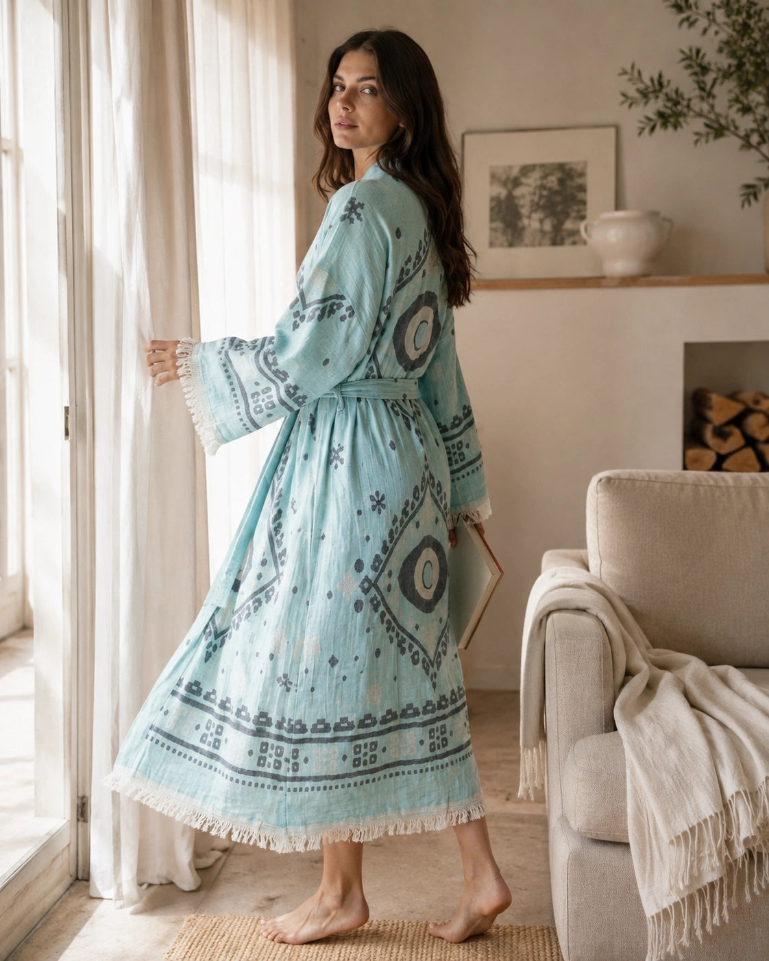 Woman wearing the blue Ocean cotton kimono robe at home, showing its long relaxed fit, evil eye pattern, belted waist and fringe trim.