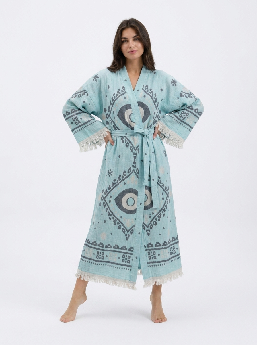 Front view of a model wearing a long aqua blue cotton kimono robe with oversized charcoal and cream evil eye motifs, geometric border details, matching waist belt, wide sleeves, and cream fringe trim.