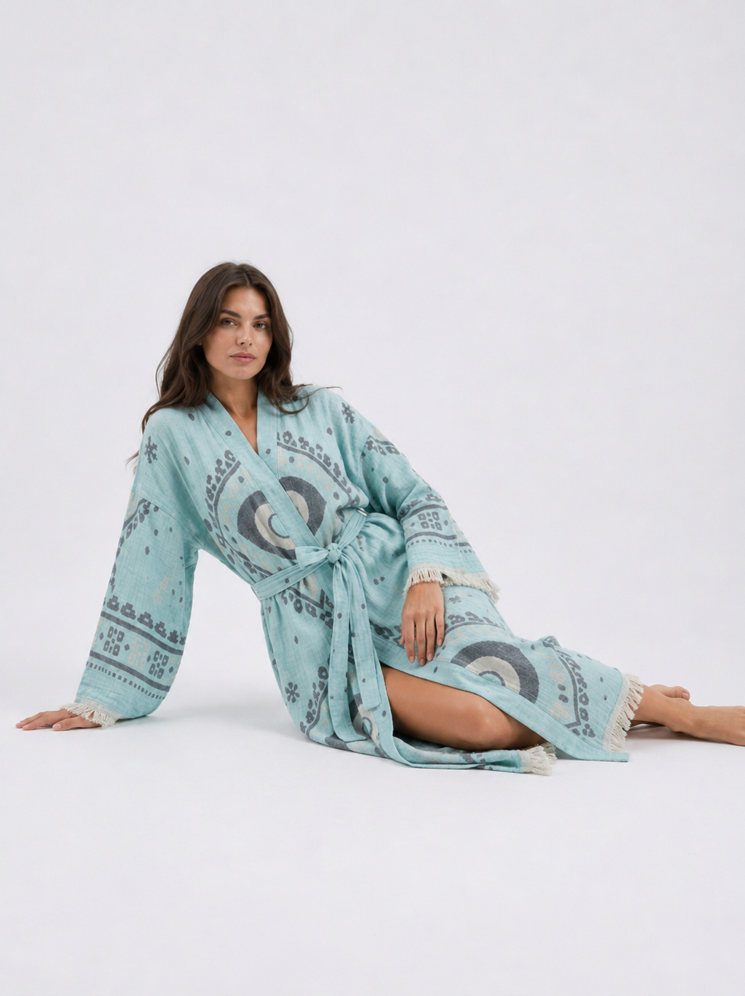 Reclining model wearing an aqua blue cotton kimono robe with oversized charcoal and cream evil eye motifs, a belted waist, wide fringed sleeves, and a relaxed full-length silhouette.