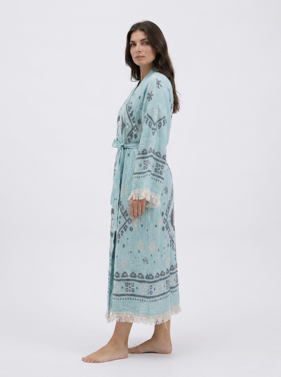 Side view of a long aqua blue cotton kimono robe showing its relaxed silhouette, oversized evil eye pattern, geometric sleeve details, matching belt, and soft cream fringe along the cuffs and hem.