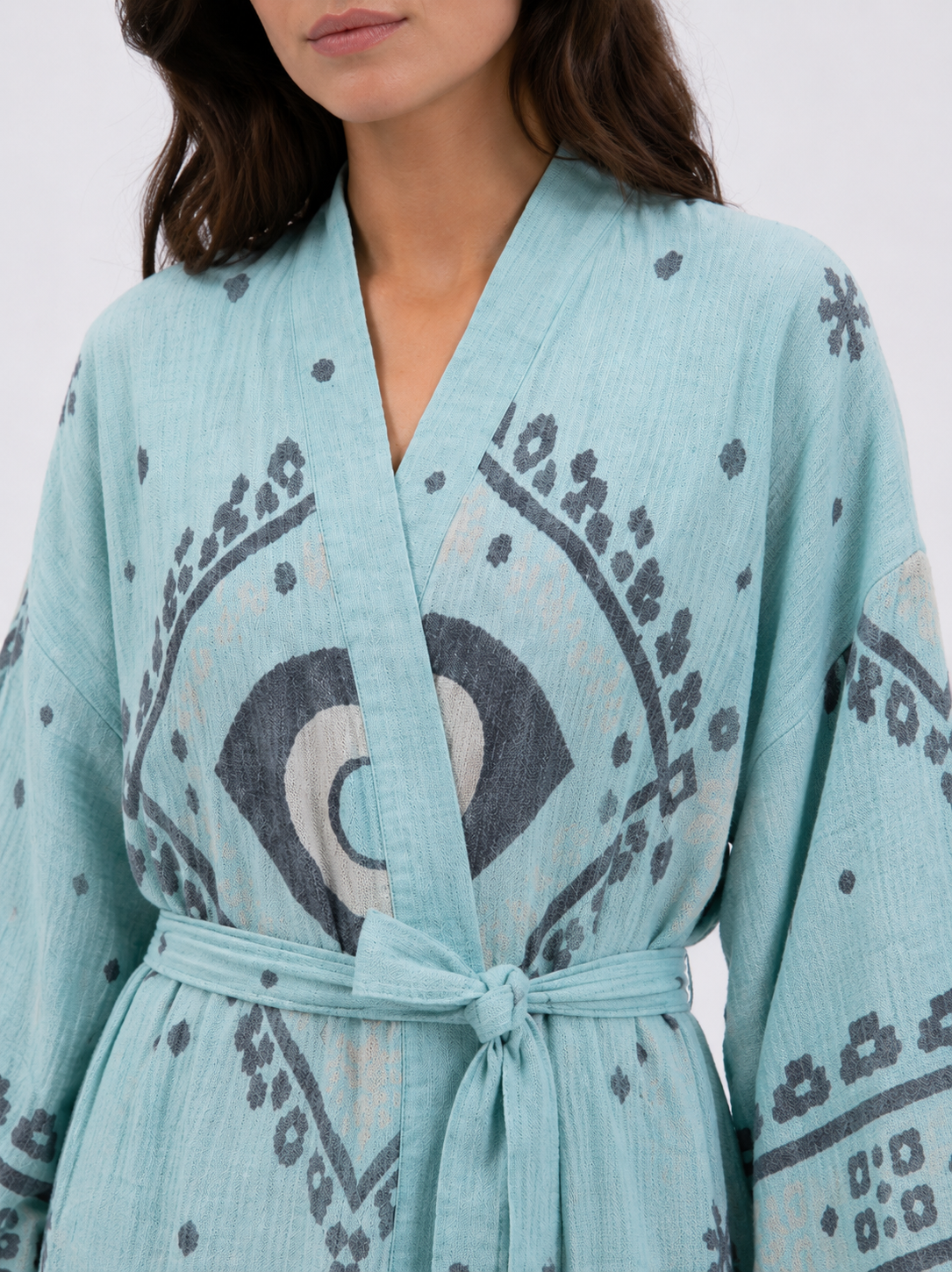 Close-up of an aqua blue cotton kimono robe showing the textured natural fabric, charcoal and cream evil eye motif, wrap neckline, matching waist tie, and geometric sleeve pattern.