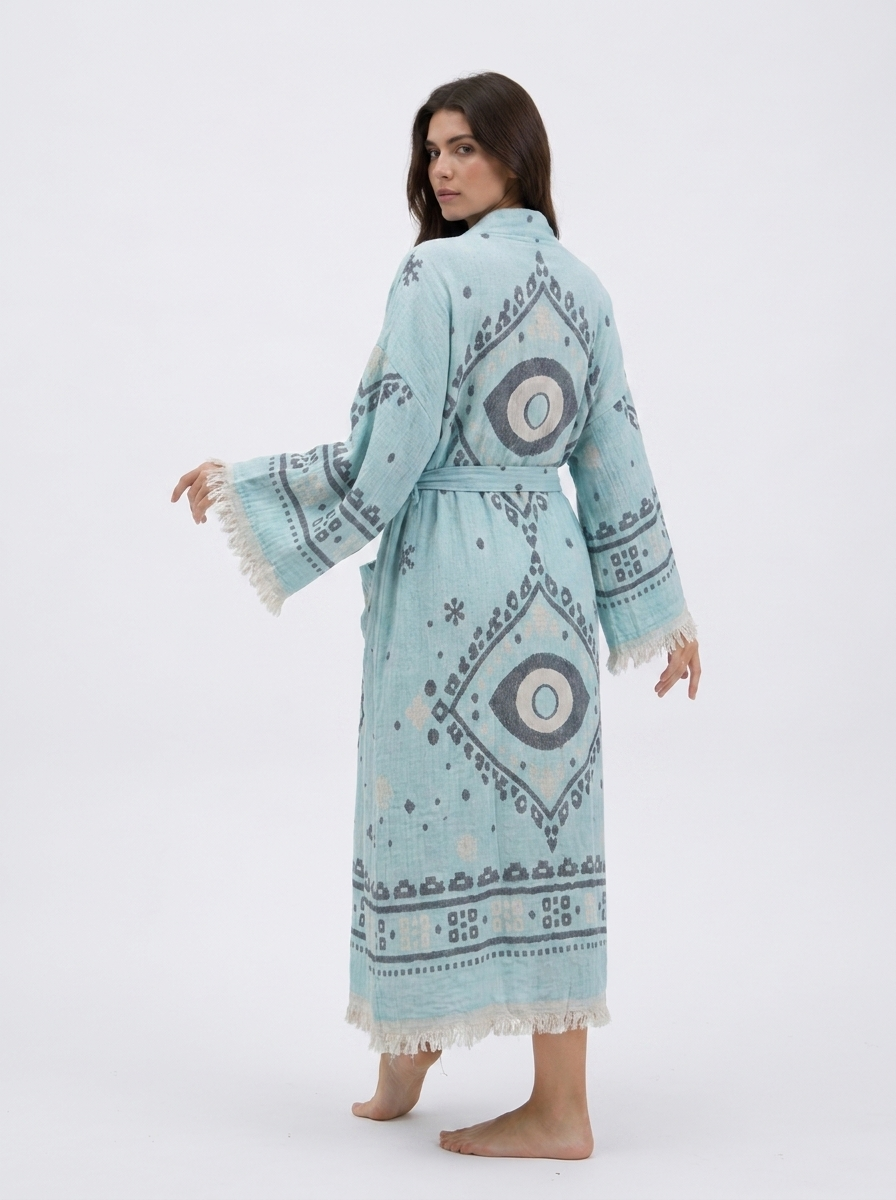 Back view of an aqua blue cotton kimono robe featuring large charcoal and cream evil eye designs, geometric woven-style motifs, a matching tie belt, wide fringed sleeves, and an ankle-length hem.