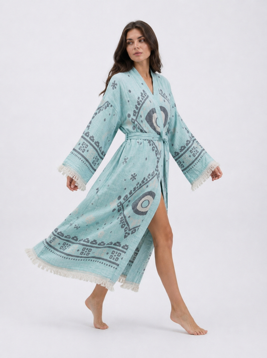 Model walking in a flowing aqua blue cotton kimono robe with charcoal evil eye motifs, wide fringed sleeves, matching waist belt, side opening, and relaxed ankle-length fit.