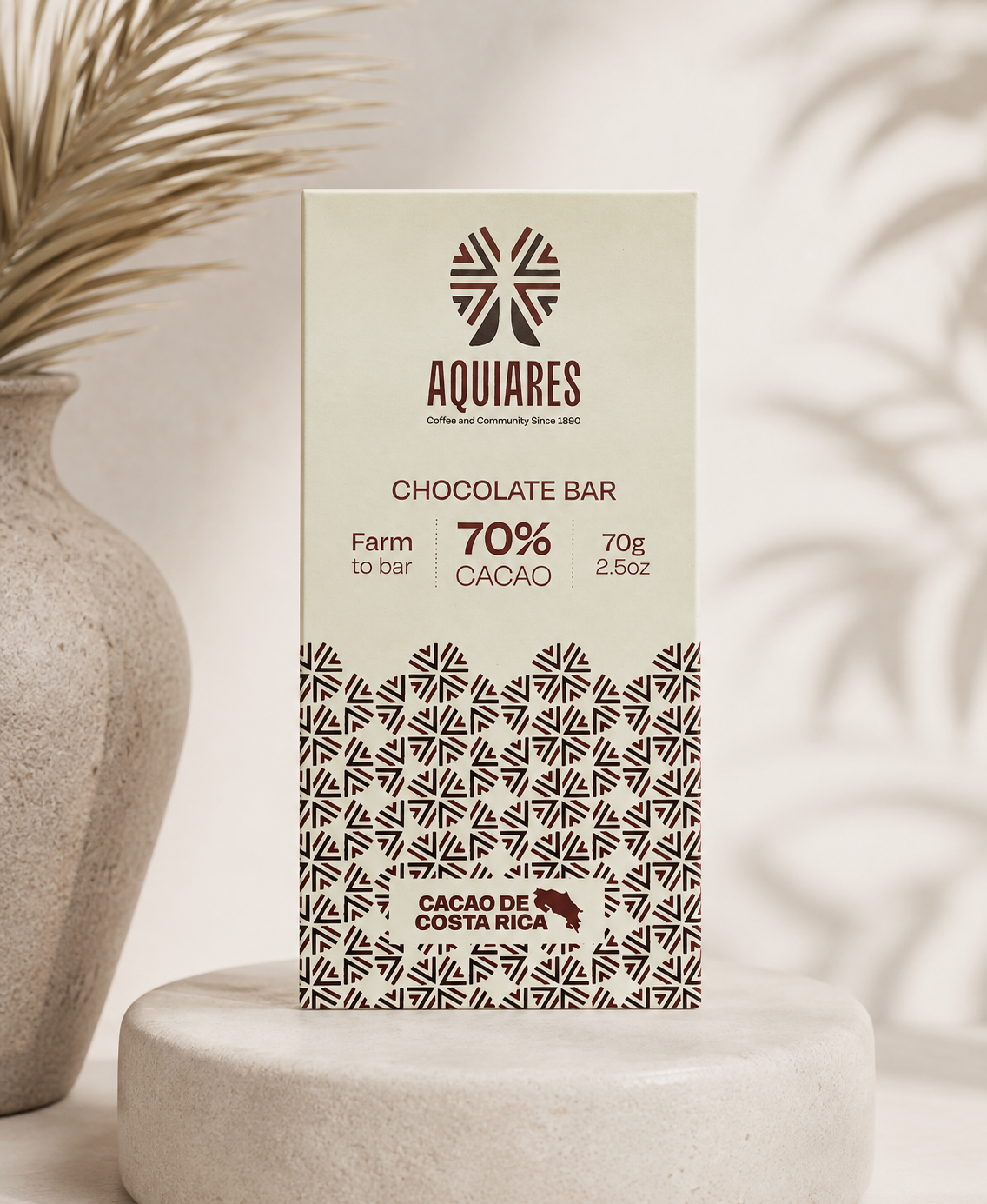 A rectangular Aquiares chocolate bar package standing upright on a round pedestal, with a beige background and dark brown text and pattern. The package reads 'AQUIARES', 'CHOCOLATE BAR', 'Farm to bar 70% CACAO', and 'CAGAO DE COSTA RICA'.