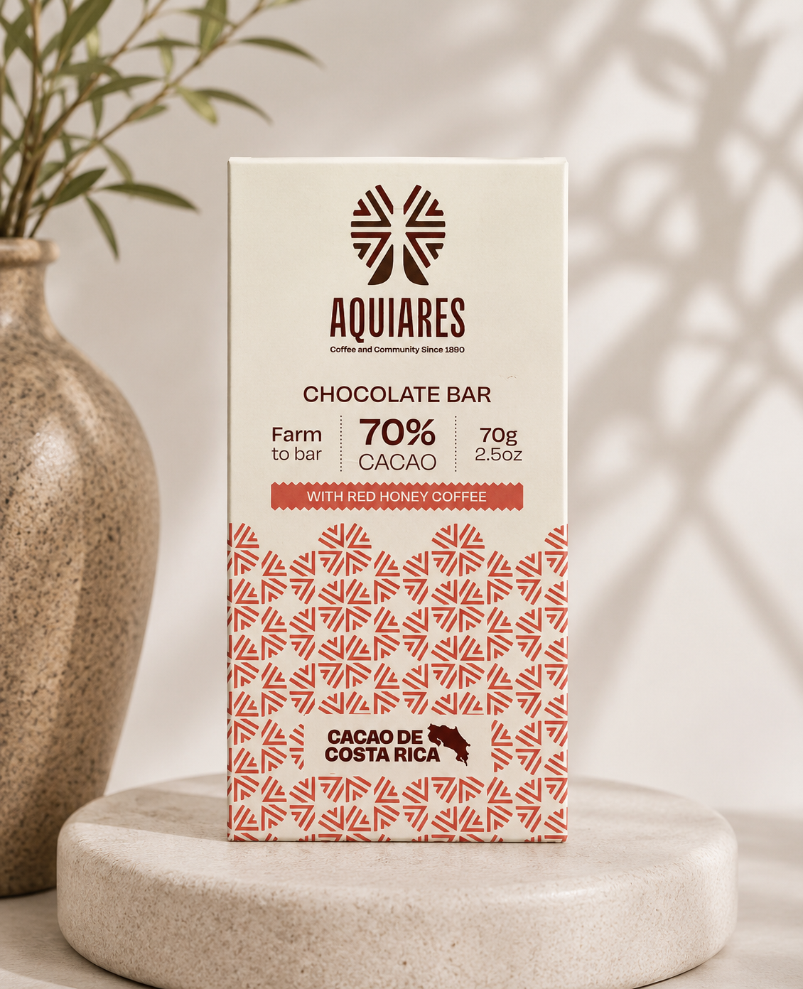 A rectangular box of Aquiares chocolate bar stands upright on a round stone pedestal. The packaging is off-white with dark brown text and a coral-red patterned lower section.