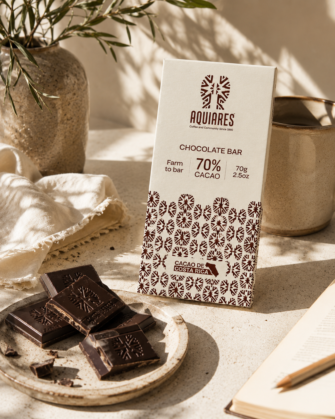 A boxed chocolate bar labeled AQUIARES 70% CACAO stands on a tabletop beside several broken dark chocolate pieces. The scene includes a ceramic vase with greenery, a folded cloth, a metal cup, and an open book in warm natural light.
