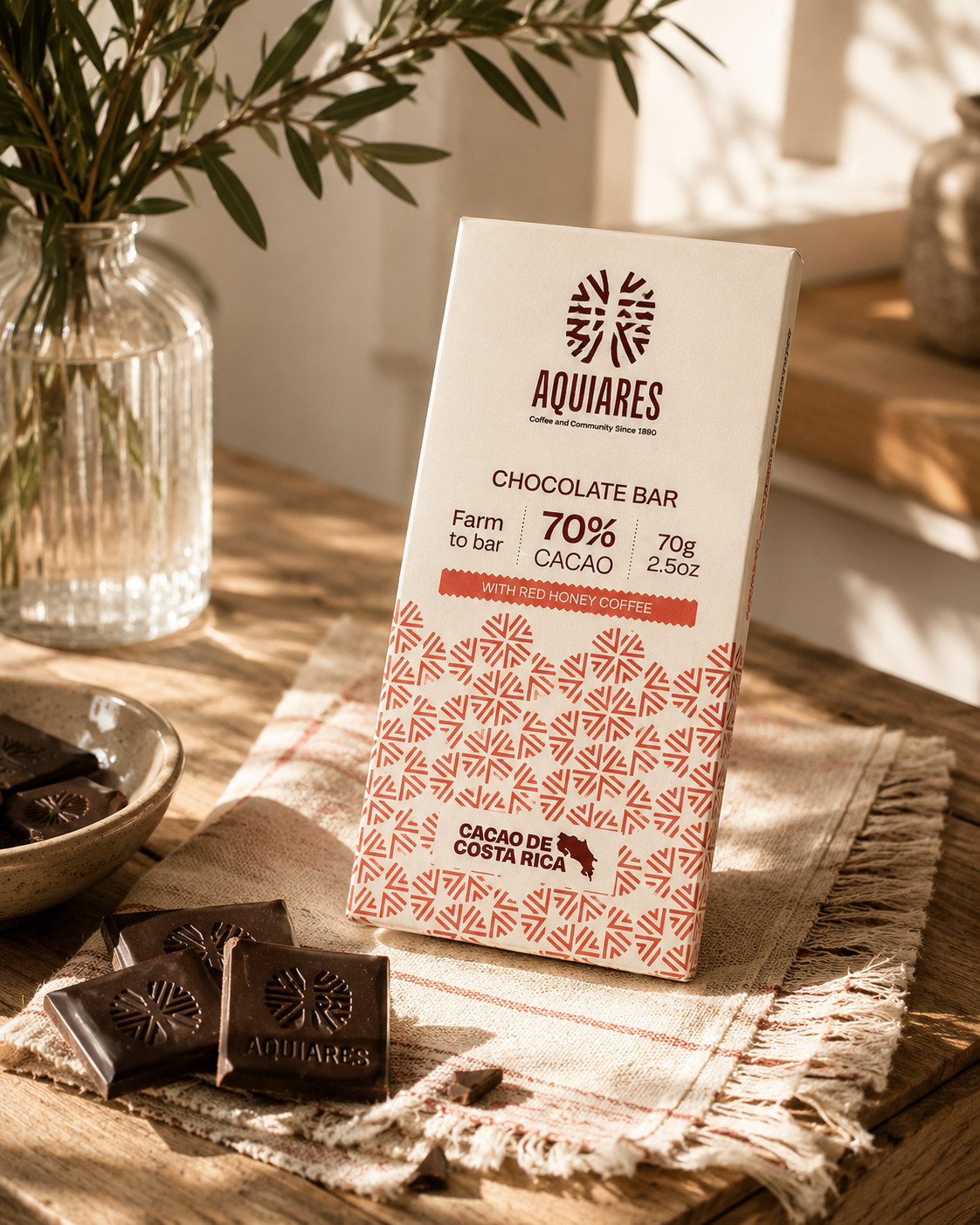 A package of Aquiareas 70% cacao chocolate bar standing on a rustic woven cloth on a wooden table, with chocolate pieces and a glass vase in the background.