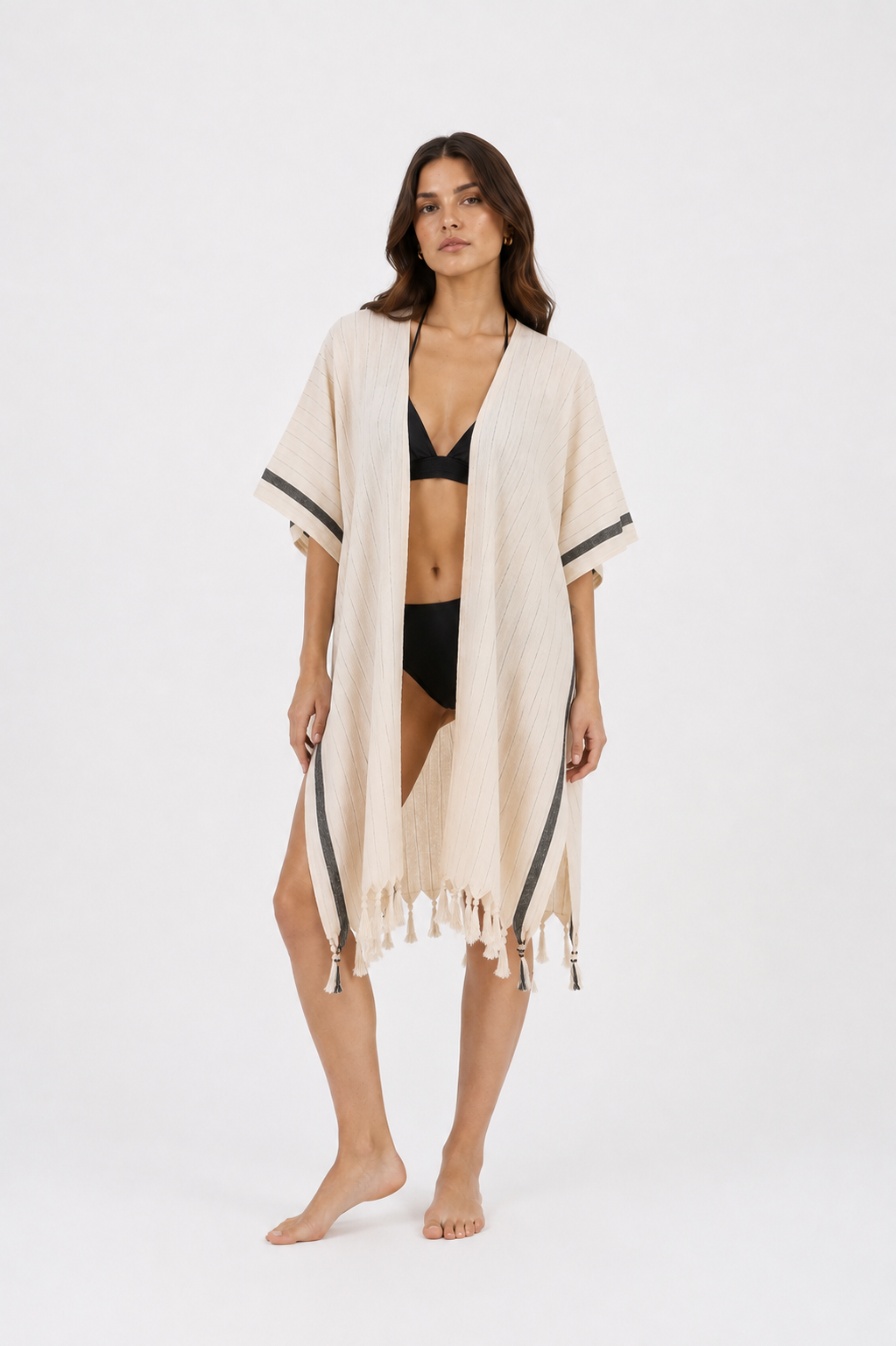 A woman stands barefoot against a plain white studio background wearing a light beige open-front cover-up with black trim and fringe, over a black bikini.