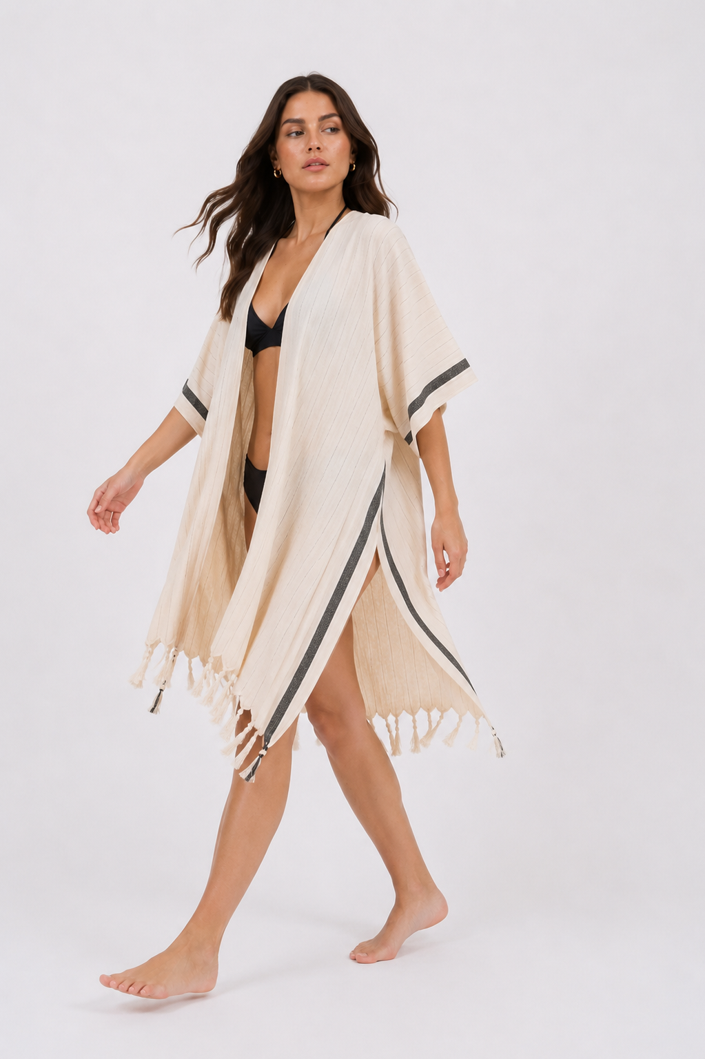 A woman modeling a lightweight beige open-front Asya cover-up with black trim and fringe, worn over a black bikini against a plain light gray studio background.