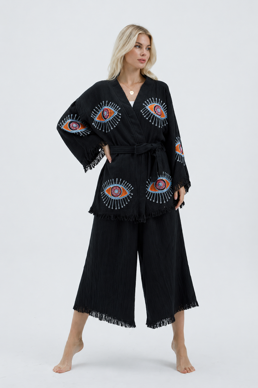 Front view of a model wearing a black textured cotton kimono jacket and matching wide-leg pants set, decorated with colorful orange, red, blue, and white evil eye motifs, a wrap front, waist belt, wide sleeves, and fringed hems.