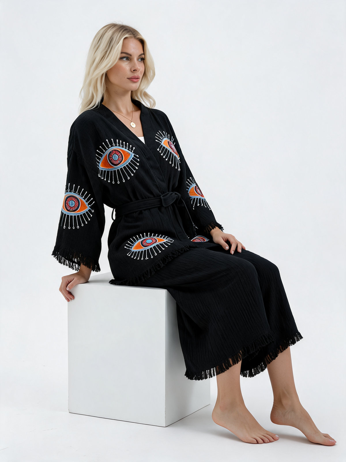 Seated model wearing a black textured cotton kimono jacket and matching wide-leg pants, featuring colorful evil eye motifs, a relaxed wrap silhouette, matching belt, and fringed sleeve and pant hems.