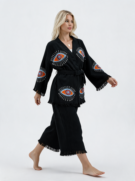 Model walking in a black cotton kimono jacket and cropped wide-leg pants set with colorful evil eye designs, flowing wide sleeves, a belted waist, and fringe-trimmed edges.