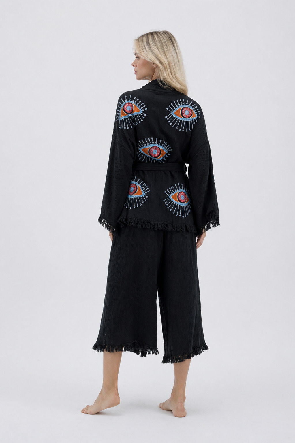 Back view of a black cotton kimono and wide-leg pants set featuring colorful evil eye motifs across the jacket, a matching fabric belt, relaxed wide sleeves, cropped pants, and soft fringe trim.