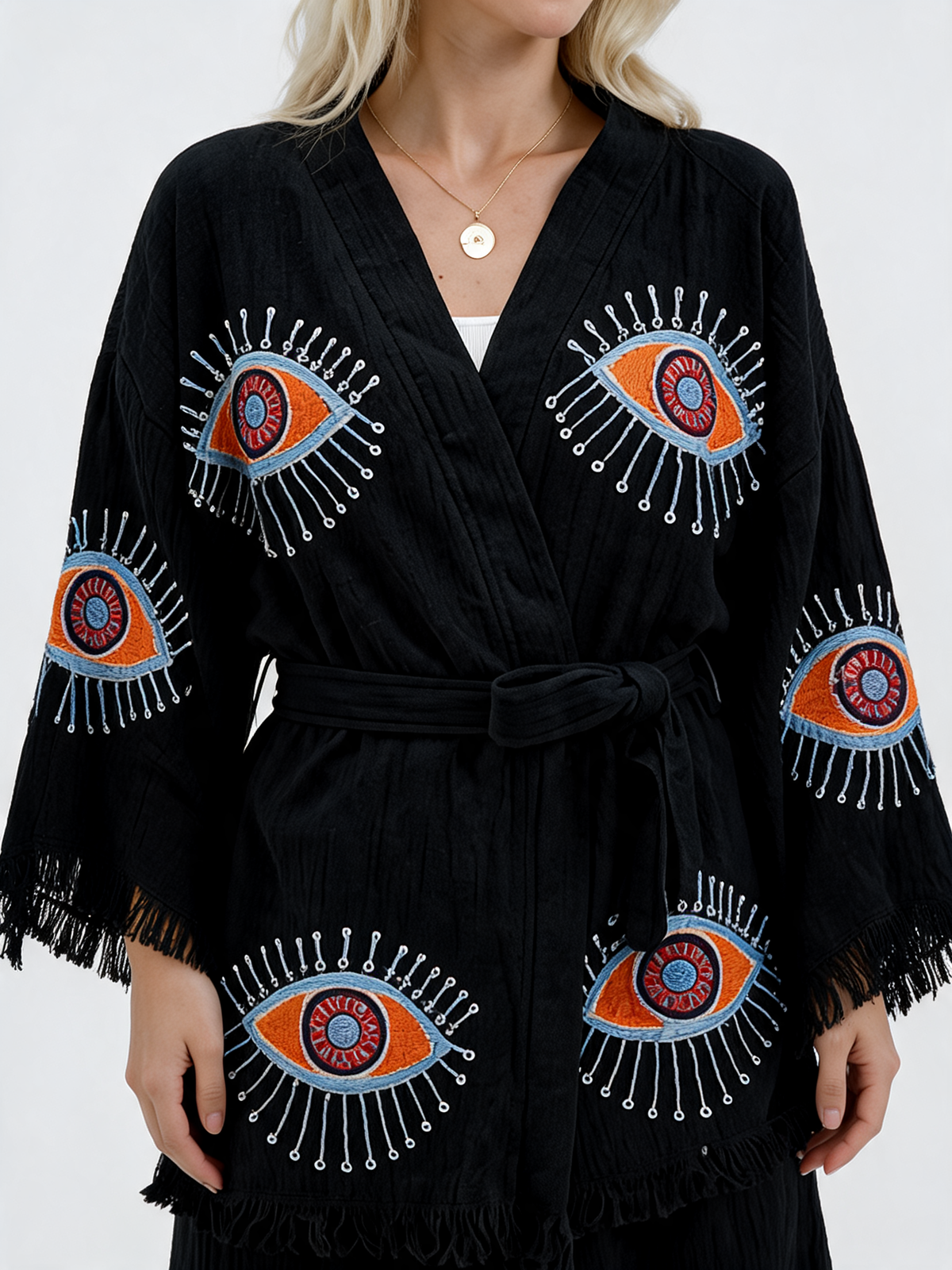 Close-up of a black textured cotton kimono jacket showing colorful evil eye embroidery in orange, red, pale blue, and white, with a wrap neckline, matching waist tie, wide sleeves, and fringe edging.