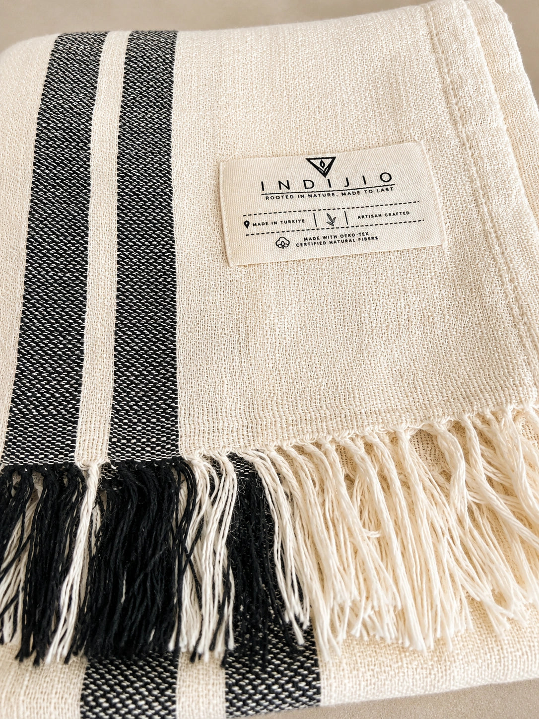 Close-up of Celine cotton towel with Indijio label, natural ivory woven texture, black stripes and hand-finished fringe.