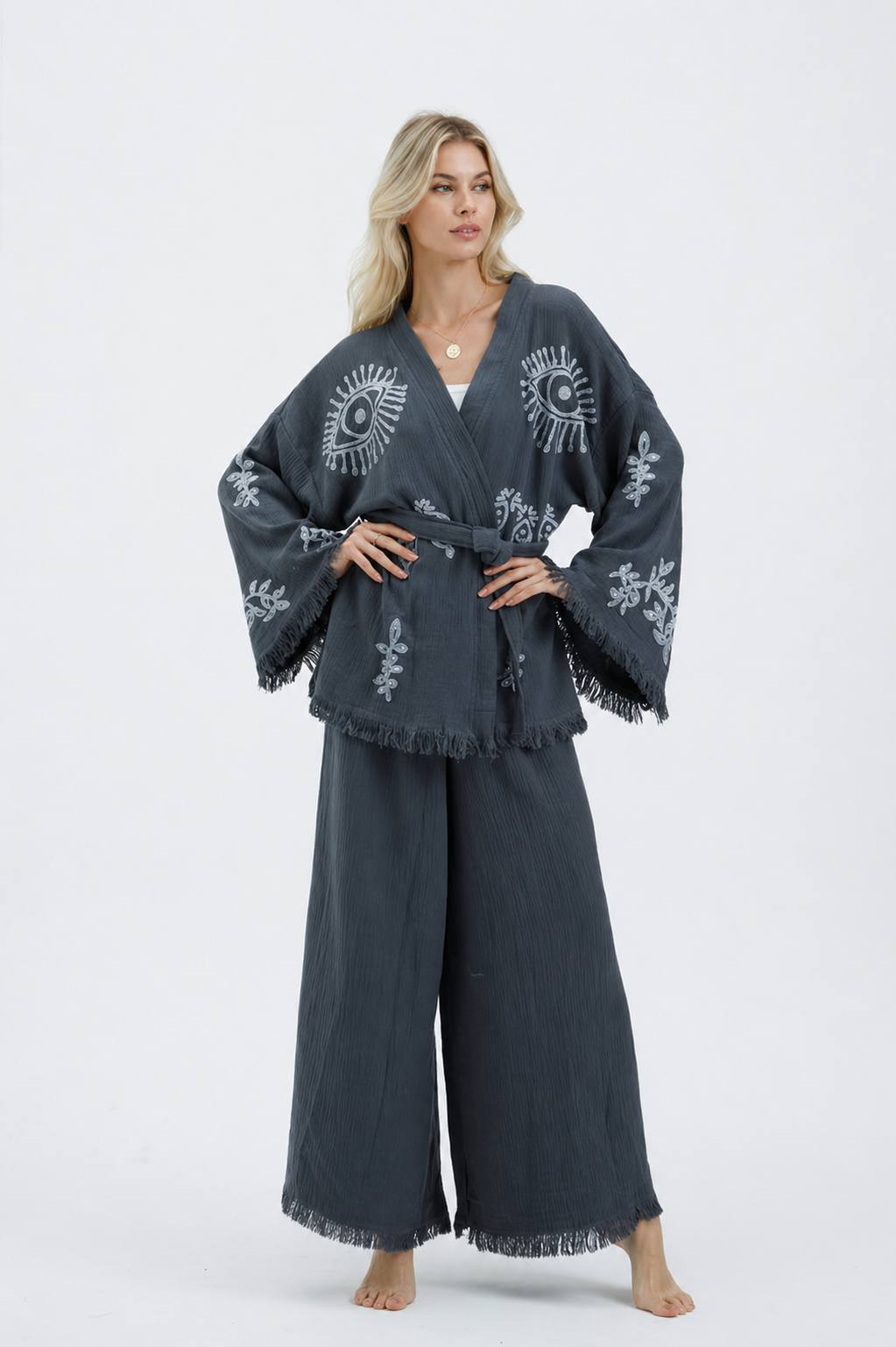 Front view of a model wearing a charcoal grey textured cotton kimono jacket and wide-leg pants set with silver evil eye and botanical motifs, a matching waist belt, wide fringed sleeves, and cropped fringe-trim pants.