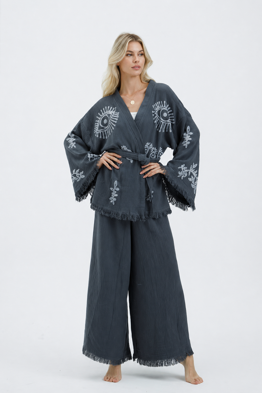 Front view of a model wearing a charcoal grey textured cotton kimono jacket and wide-leg pants set with silver evil eye and botanical motifs, a matching waist belt, wide fringed sleeves, and cropped fringe-trim pants.
