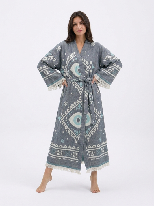 Front view of a model wearing a long charcoal grey cotton kimono robe with oversized white and aqua evil eye motifs, geometric border details, a matching waist belt, wide sleeves, and cream fringe trim.