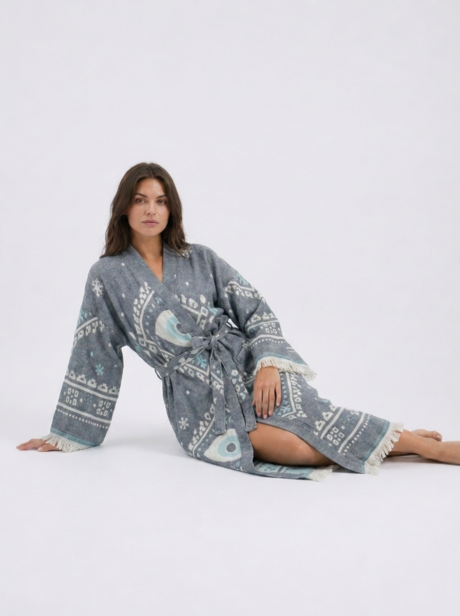 Reclining model wearing a charcoal grey cotton kimono robe with oversized white and aqua evil eye motifs, a belted waist, wide fringed sleeves, and a relaxed full-length silhouette.
