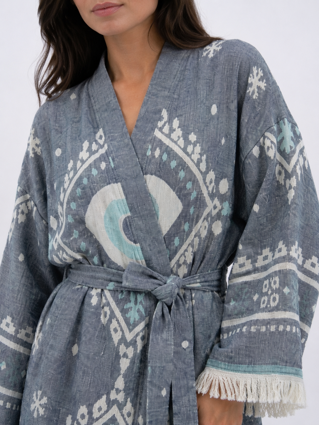 Close-up of a charcoal grey cotton kimono robe showing the textured fabric, white and aqua evil eye motif, wrap neckline, matching waist belt, geometric sleeve pattern, and cream fringe edging.