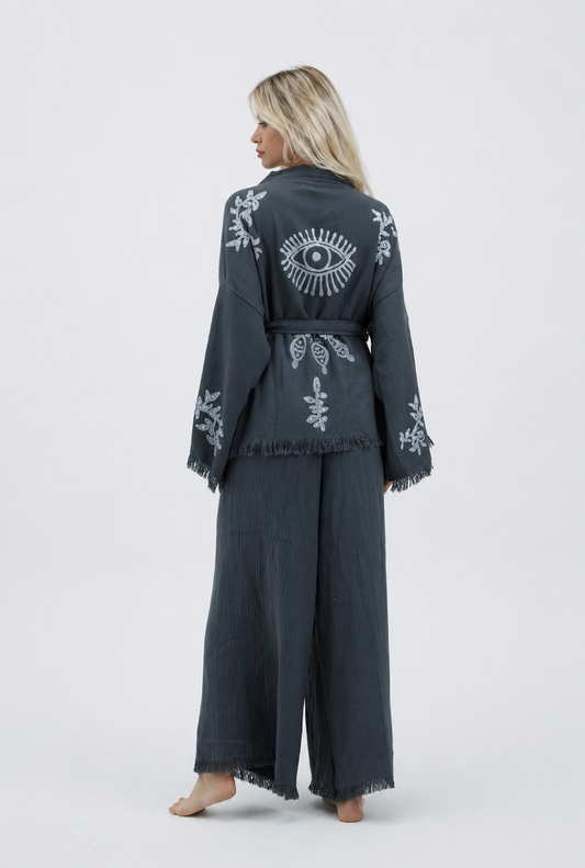 Back view of a charcoal grey cotton kimono and wide-leg pants set featuring a large silver evil eye motif, delicate botanical designs, a matching tie belt, wide sleeves, and fringed hems.