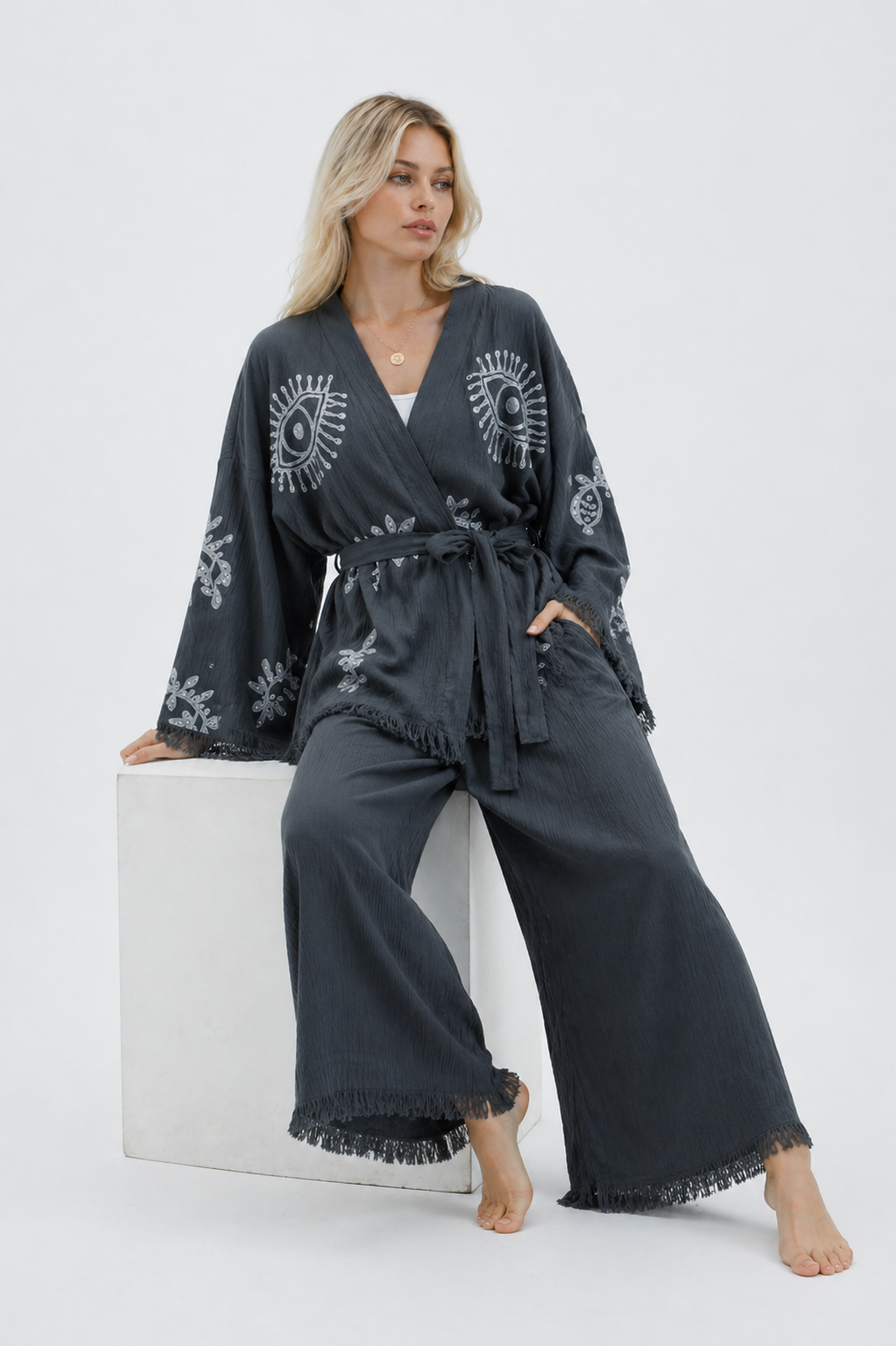 Seated model wearing a charcoal grey cotton kimono jacket and wide-leg pants set with silver evil eye and botanical patterns, a belted waist, wide fringed sleeves, and relaxed cropped pants.