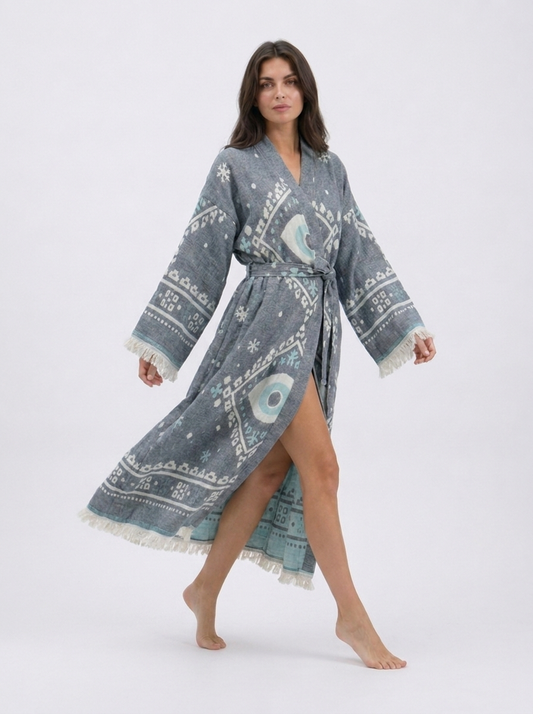 Model walking in a flowing charcoal grey cotton kimono robe with white and aqua evil eye motifs, wide fringed sleeves, a matching waist belt, side opening, and ankle-length hem.