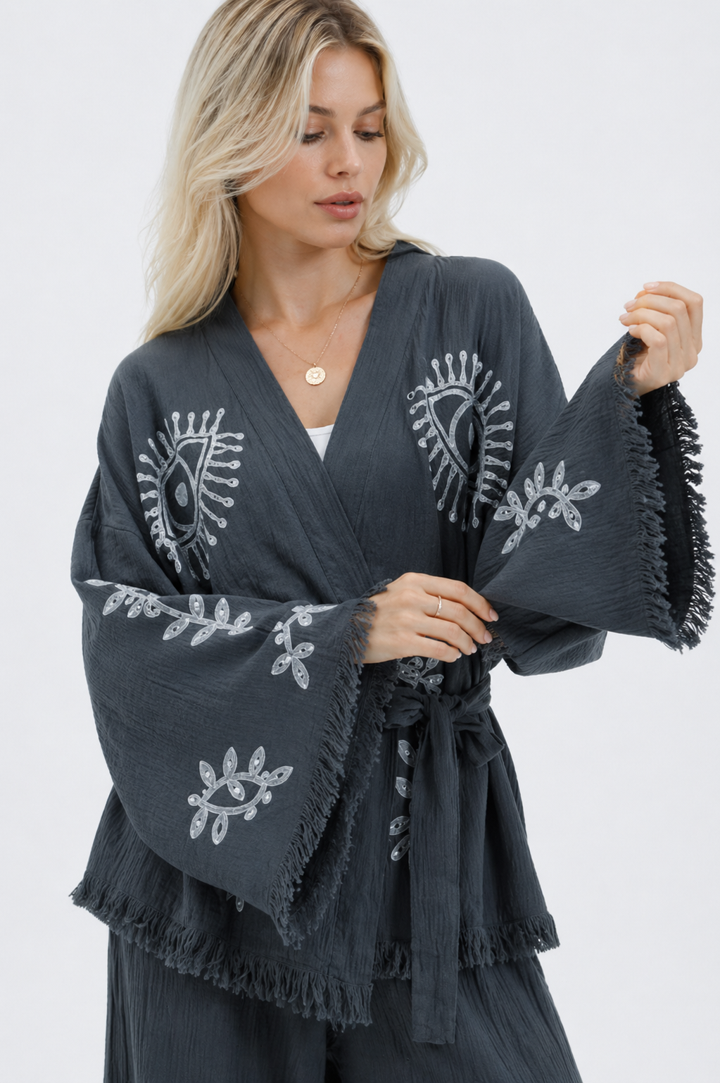 Close-up of a charcoal grey textured cotton kimono jacket showing silver evil eye and leafy botanical motifs, oversized sleeves, raw fringe edging, a wrap neckline, and a matching fabric belt.