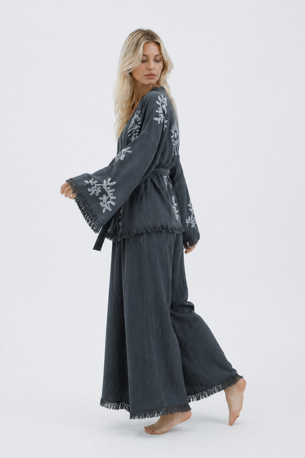 Side-back view of a model wearing a charcoal grey textured cotton kimono and wide-leg pants set, showing silver evil eye and botanical motifs, flowing sleeves, fringe trim, and relaxed movement.