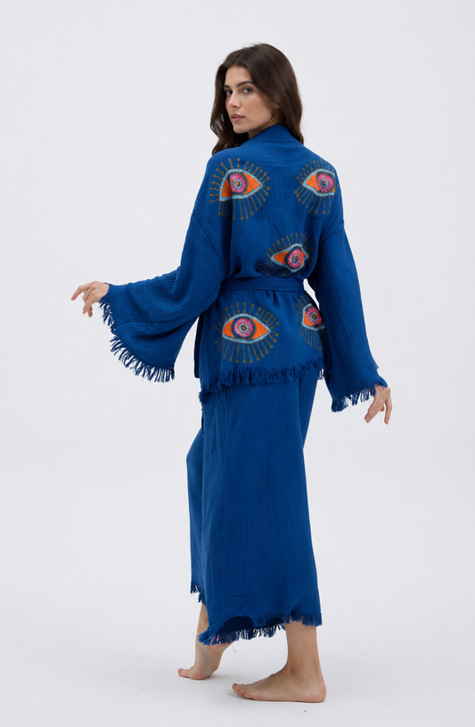 Back view of a cobalt blue cotton kimono and wide-leg pants set featuring large colorful evil eye motifs across the jacket, a matching fabric belt, flowing wide sleeves, and fringe-trimmed edges.