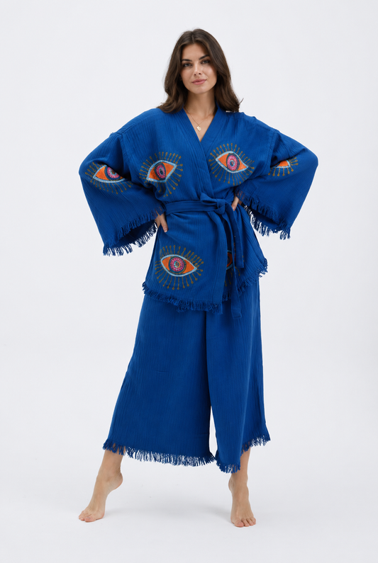 Front view of model wearing a cobalt blue cotton kimono and pants set with colorful evil eye motifs, matching belt, wide sleeves and fringe hems.