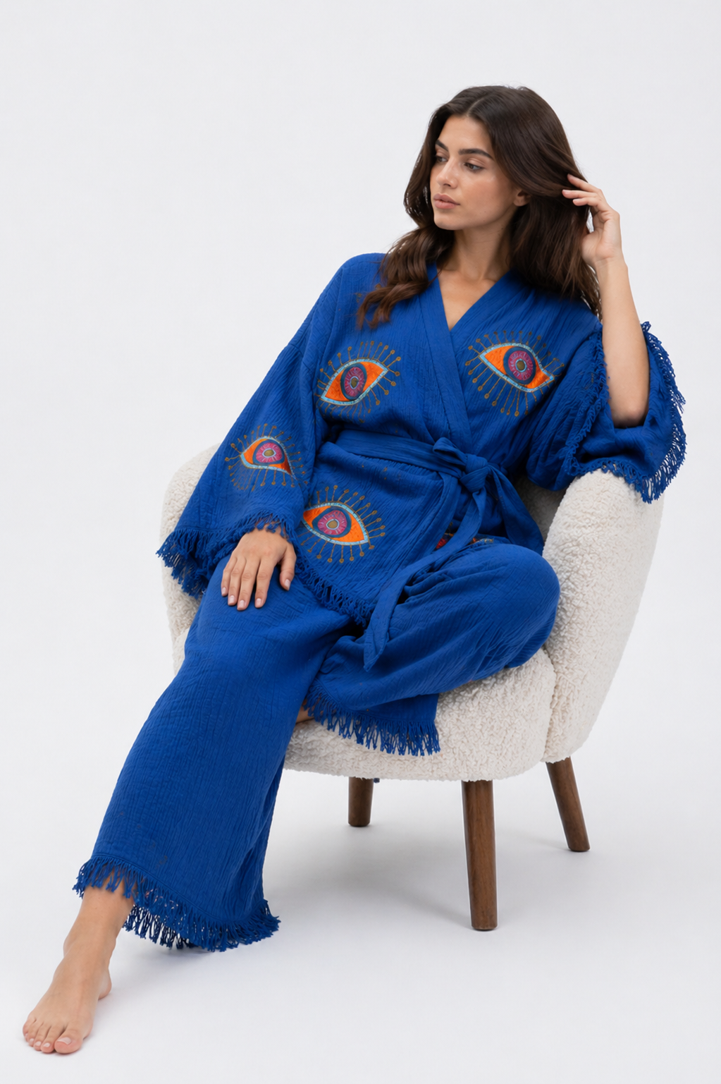 Seated model wearing a cobalt blue cotton kimono jacket and wide-leg pants set with colorful evil eye designs, a tied waist, oversized fringed sleeves, and relaxed lounge-inspired styling.
