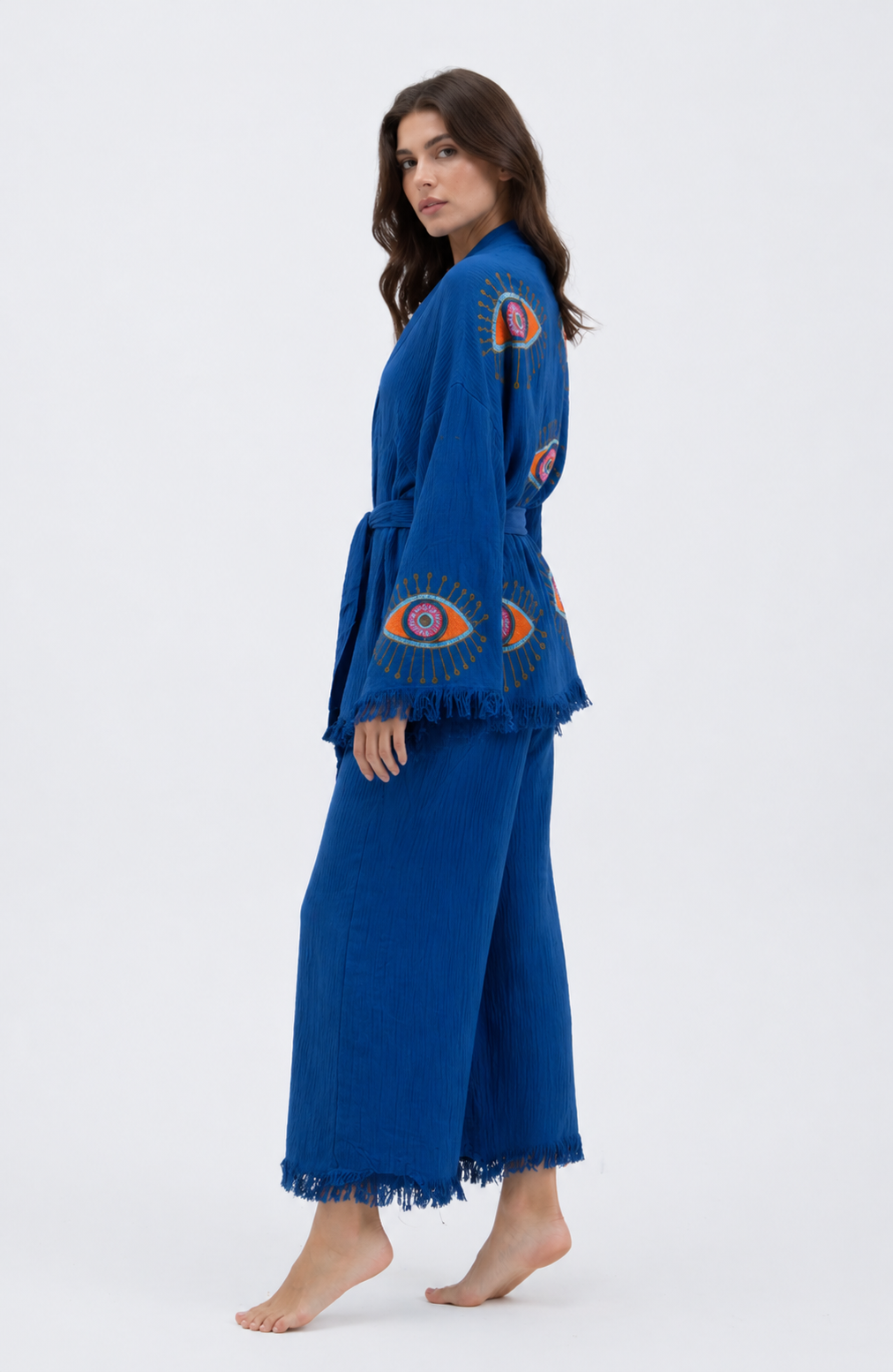 Side view of a cobalt blue textured cotton kimono jacket and wide-leg pants set showing colorful evil eye artwork, a relaxed belted silhouette, cropped wide-leg pants, and soft fringe trim.